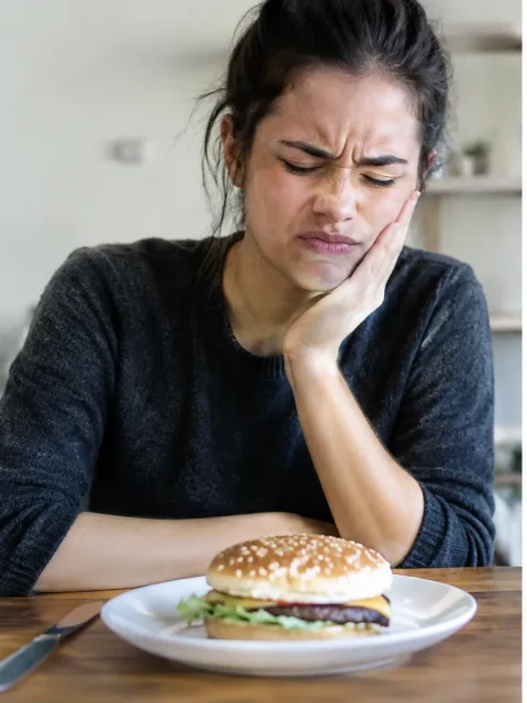 Woman wincing with jaw pain in front of a burger